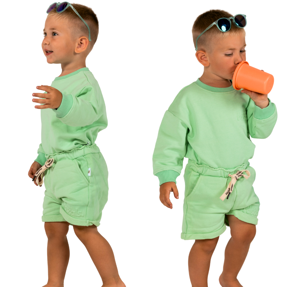 Child in mint green spill resistant outfit posing drinking a sippy cup