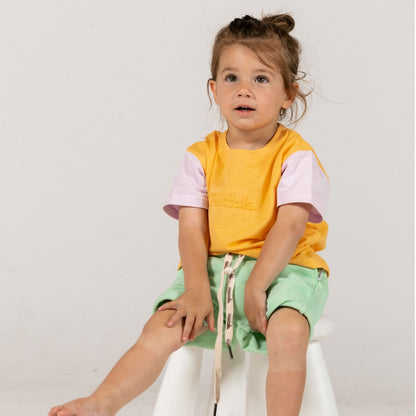 Child wearing a apricot orange and lavender spill resistant t-shirt with mint green shorts by Natural Rascals sitting on a white stool against a plain background