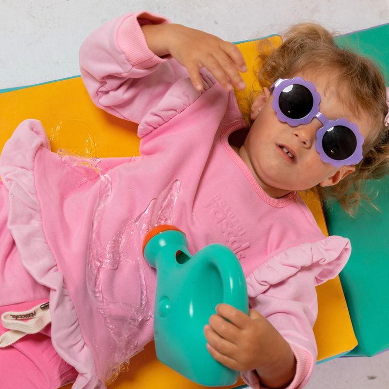 Child in raspberry pink spill resistant outfit by Natural Rascals with purple sunglasses holding a teal toy on a colorful background