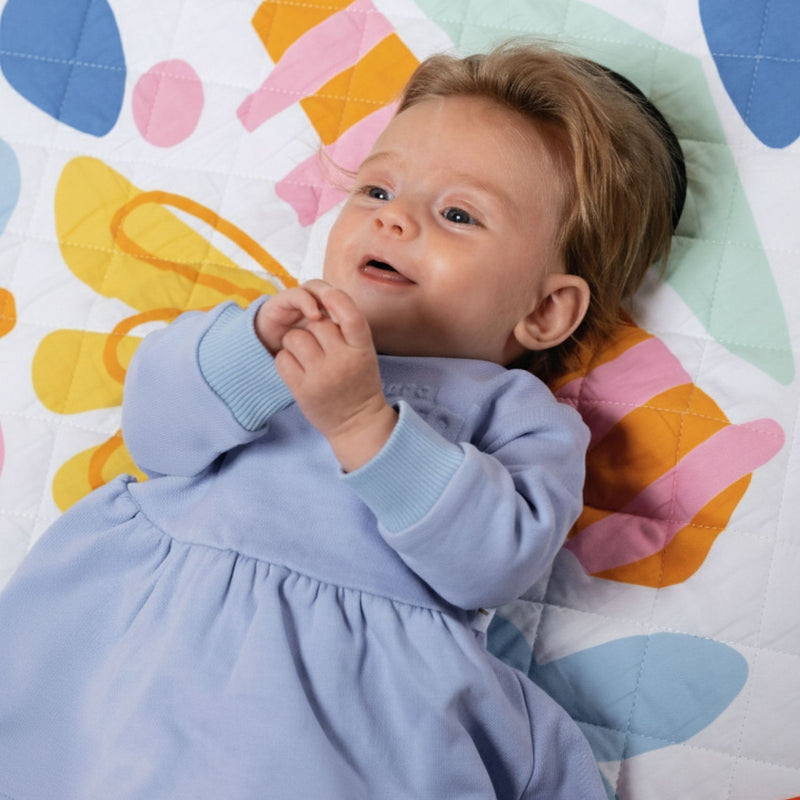 Happy baby in a periwinkle lavender purple spill resistant dress lying on a colorful quilt with butterfly design by Natural Rascals