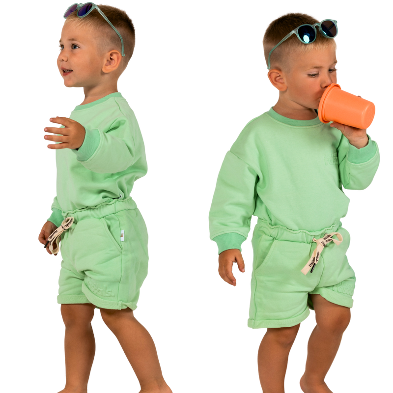 Child in mint green spill resistant outfit posing drinking a sippy cup