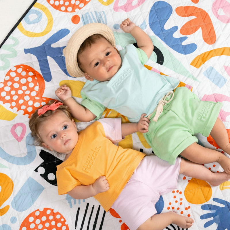 Two babies lying on a colorful play mat with abstract patterns wearing natural rascals spill resistant outfits 