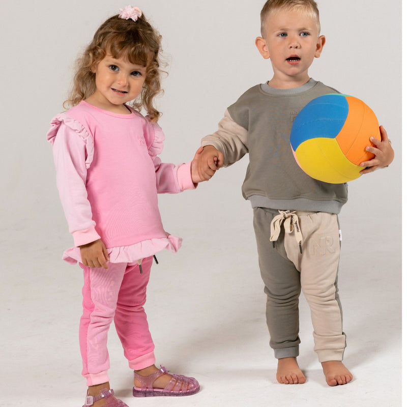 Two children, a girl in raspberry pink and a boy in earthy and oat gray with a colorful ball, standing against a plain background by natural rascals