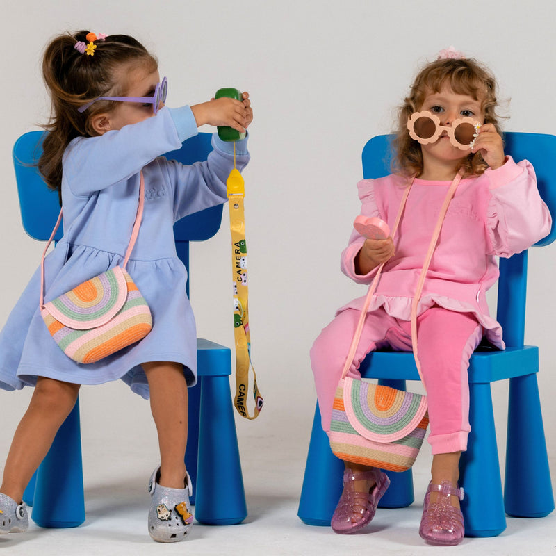 Two children sitting on blue chairs, wearing colorful sunglasses and holding rainbow bags. The children are wearing spill resistant clothing sets in lavender and raspberry and candy pink by Natural Rascals