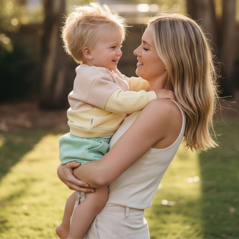 Woman holding a child in a park with trees and grass in the background. The child is wearing spill resistant lemonade yellow and oat sweat paired with mint green shorts by Natural Rascals