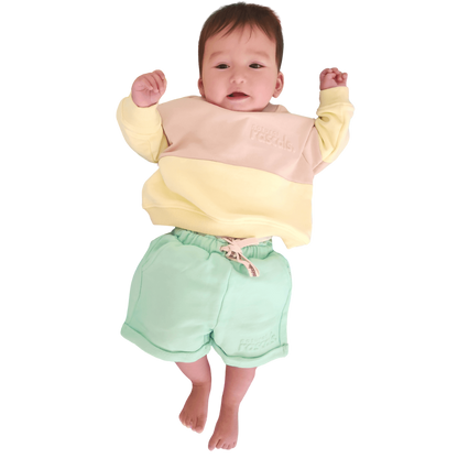 Baby wearing a lemonade yellow, oat and mint green Spill resistant shorts and sweat set by Natural Rascals on a transparent background