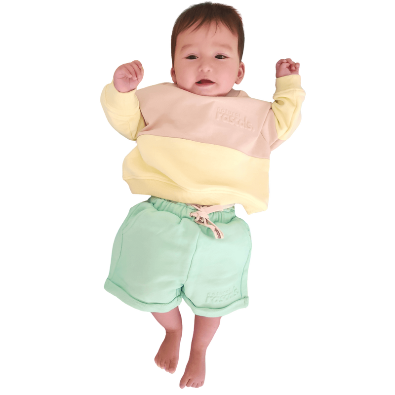 Baby wearing a lemonade yellow, oat and mint green Spill resistant shorts and sweat set by Natural Rascals on a transparent background
