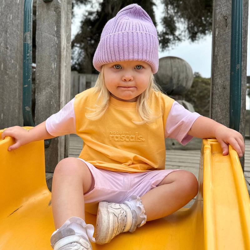 Child sitting on yellow slide in apricot orange and soft lilac spill-resistant baby toddler tee by Natural Rascals.